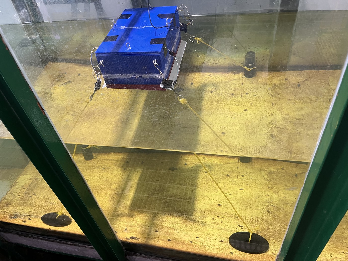 Model floating PV tipe barge dengan empat tali tambat saat diuji di wave flume BRIN Yogyakarta, Mei 2026, dilihat dari balik dinding kaca